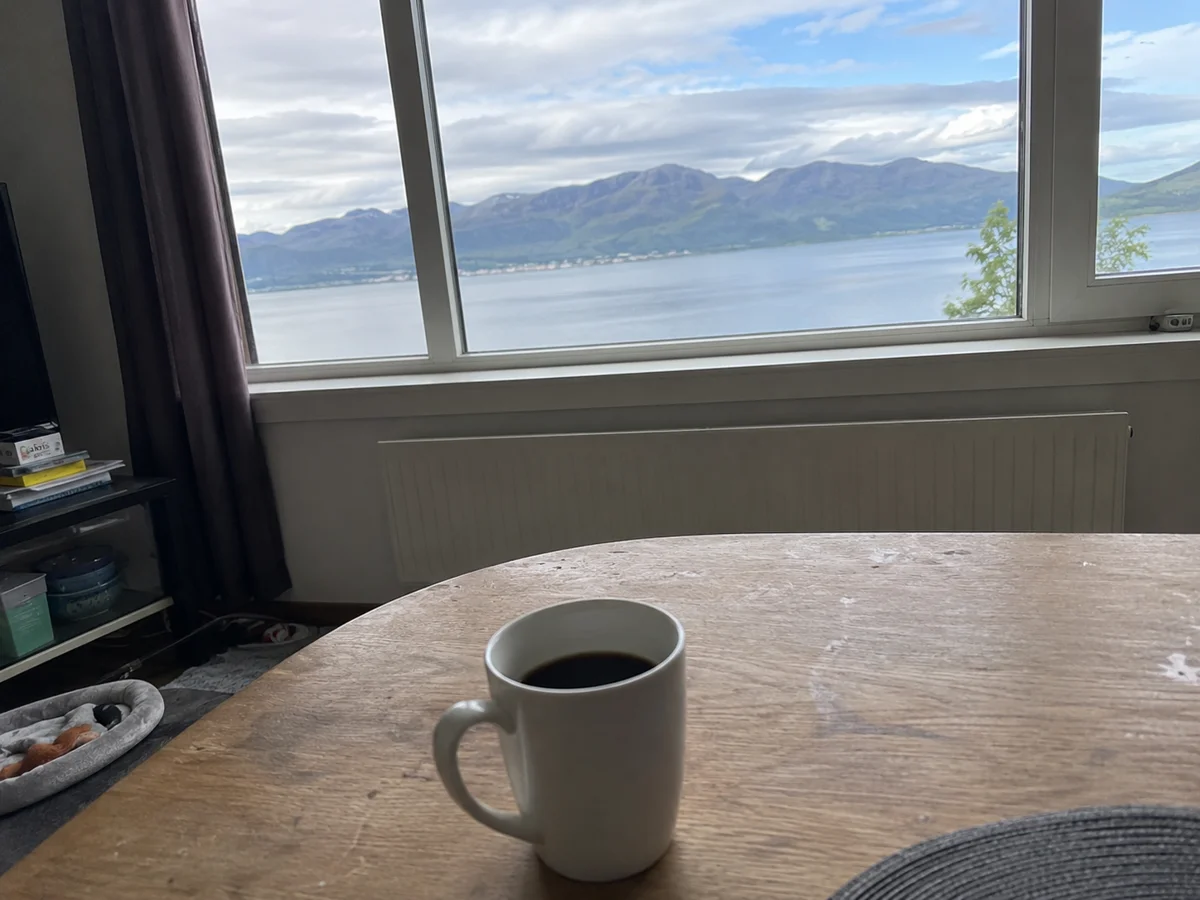 KI-generert testbilde av en kaffekopp på et trebord foran et vindu med utsikt over fjord og fjell, laget med ChatGPT